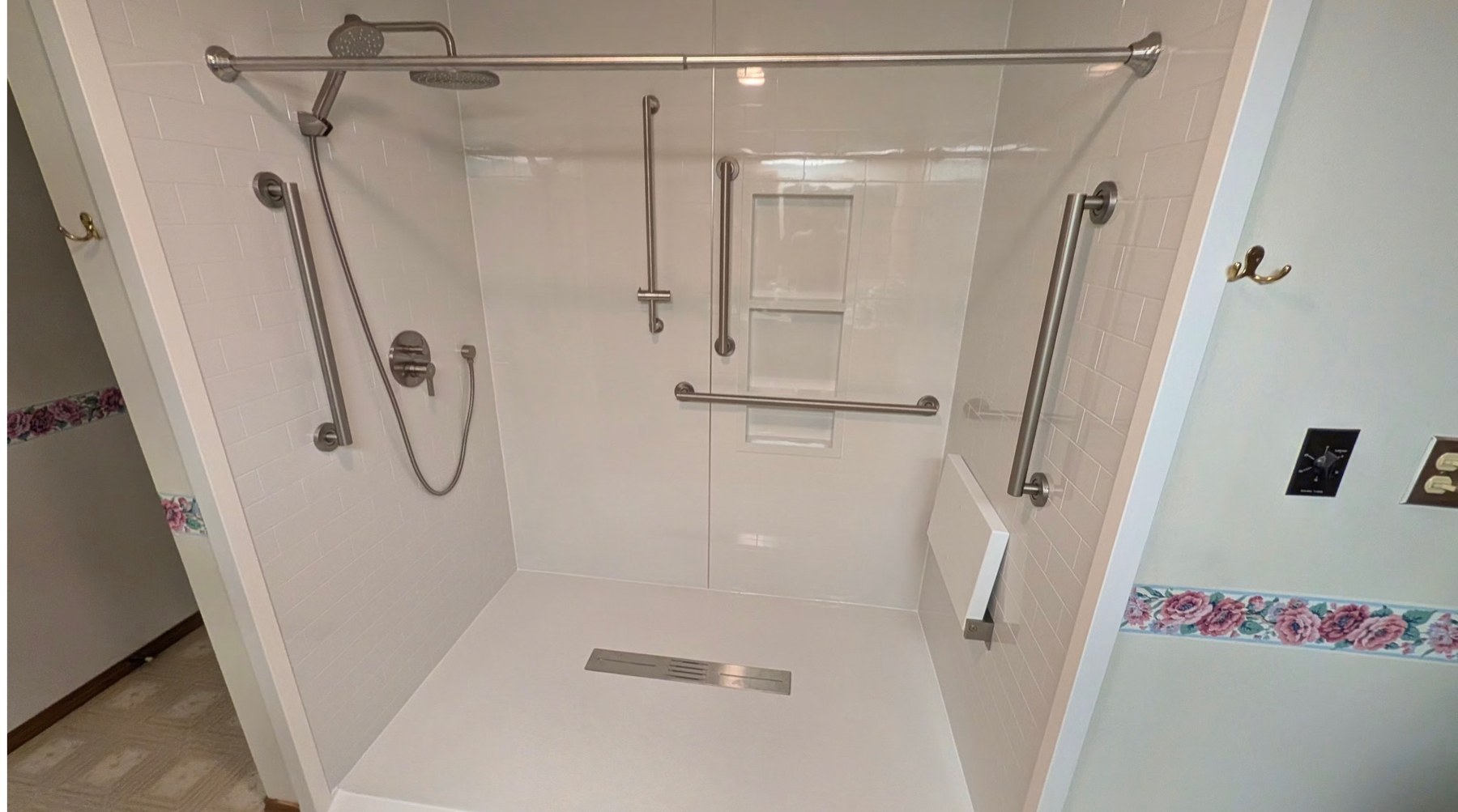 Roll in walk in shower with white subway look walls, a brushed nickel rain shower head and handheld, grab bars on three walls, a recessed niche, a white fold down seat and a linear drain installed by American Shower Company in a Wisconsin home